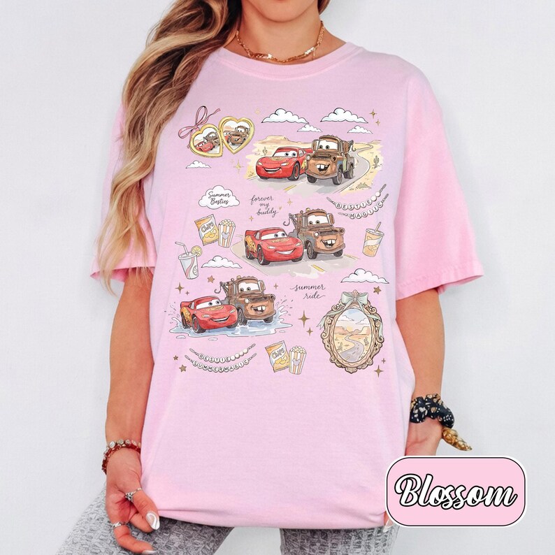 Puede incluir: Camiseta rosa claro con dibujos de coches de dibujos animados y los textos "Summer Buddies", "Forever my buddy" y "Summer ride". El dise&ntilde;o incluye nubes, estrellas y un paisaje des&eacute;rtico. La palabra "Blossom" est&aacute; en un rect&aacute;ngulo rosa.
