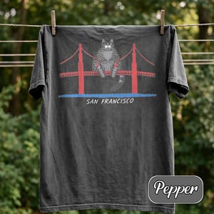 May include: Dark gray t-shirt featuring a cartoon cat sitting on the Golden Gate Bridge in San Francisco. The bridge is red with red cables, and the text "SAN FRANCISCO" is below the bridge. The shirt is hanging on a clothesline.