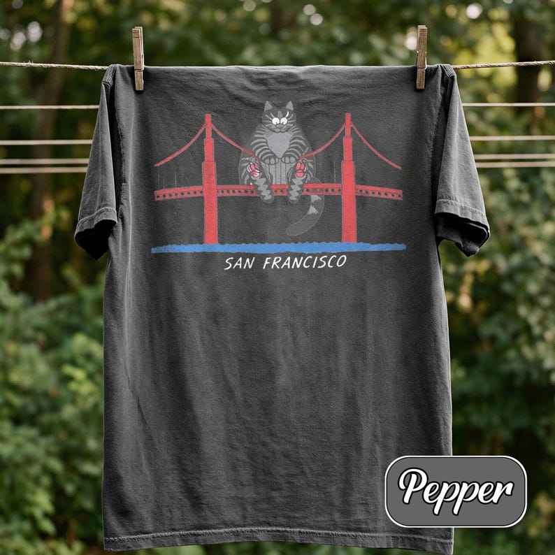 May include: Dark gray t-shirt featuring a cartoon cat sitting on the Golden Gate Bridge in San Francisco. The bridge is red with red cables, and the text "SAN FRANCISCO" is below the bridge. The shirt is hanging on a clothesline.