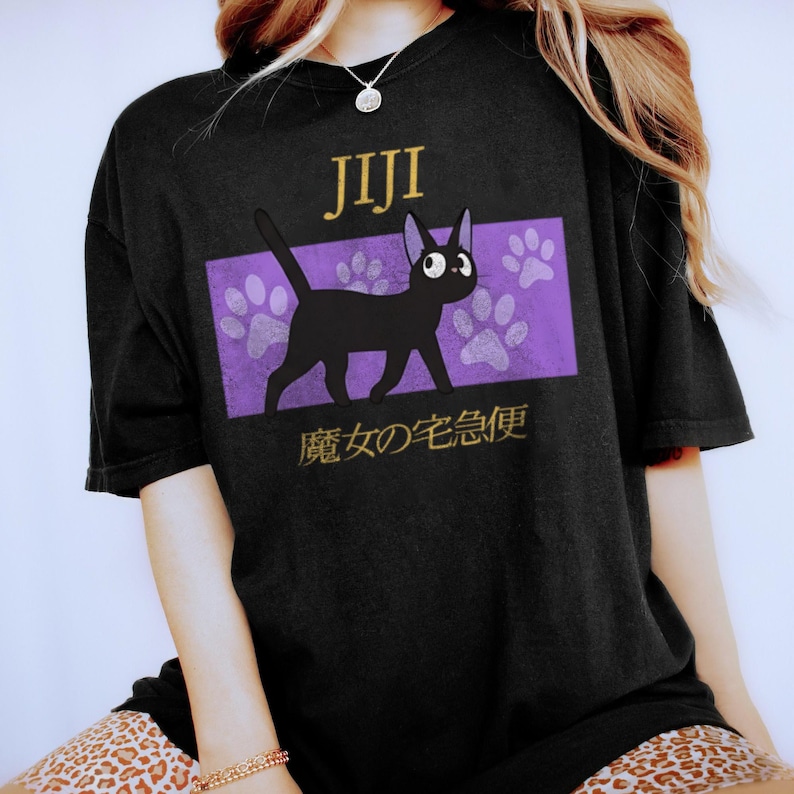 May include: A black short-sleeved t-shirt featuring a graphic of a black cat. Above the cat is the text Jiji, and below is Japanese text. The cat is positioned over a purple rectangular background with light purple paw print patterns.