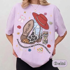 Pu&ograve; includere: Una t-shirt a maniche corte color viola chiaro con una stampa grafica raffigurante uno stivale da cowboy maculato, un cappello rosso, il nome Taylor, il numero 13, blocchi impilati con lettere, piccoli fiori e stelle.