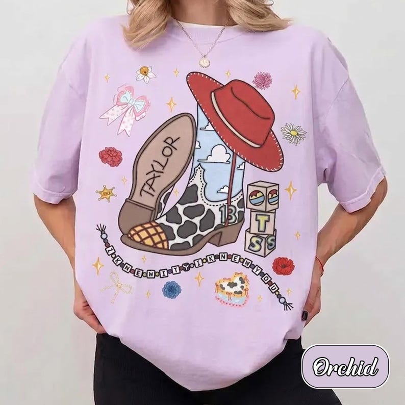 Pu&ograve; includere: Una t-shirt a maniche corte color viola chiaro con una stampa grafica raffigurante uno stivale da cowboy maculato, un cappello rosso, il nome Taylor, il numero 13, blocchi impilati con lettere, piccoli fiori e stelle.