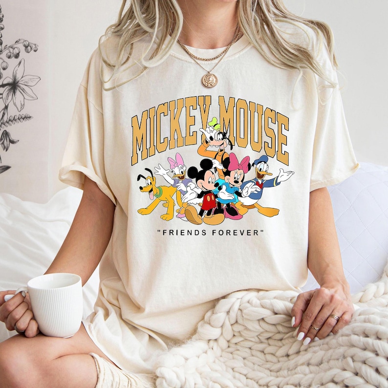 May include: Cream-colored t-shirt featuring a graphic of Mickey Mouse and friends, including Donald Duck, Goofy, and Minnie Mouse. The text "MICKEY MOUSE" is arched above the characters, with "FRIENDS FOREVER" below.