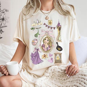 May include: Cream-colored t-shirt featuring a collection of cartoon illustrations. The design includes a portrait of a blonde woman, a purple dress, a frying pan, a tower, and other related elements. The shirt is a comfortable, casual style.
