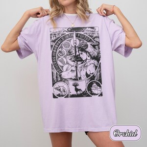 以下が含まれることがあります： 薄紫色のTシャツで、槍を持つキャラクターの黒いグラフィックプリントが施されています。周囲には森の生き物の円形イラストと「Princess Mononoke in the Forest」というテキストが描かれています。クルーネックで半袖のデザインです。