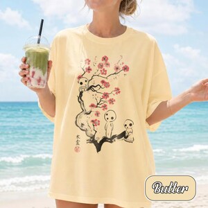 May include: A pale yellow t-shirt featuring a black ink-style illustration of a tree with pink blossoms and three small white forest spirit figures. The design includes vertical Japanese text and a red circular stamp on the lower left side.