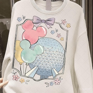 May include: Cream-colored sweatshirt featuring a colorful graphic of Mickey Mouse balloons, a geometric sphere, and a purple bow. The design includes pastel pink, yellow, and blue elements, with floral accents. The sweatshirt has a ribbed crew neck.
