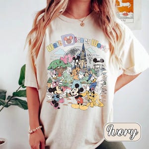 May include: Ivory-colored t-shirt featuring a colorful graphic of Disney characters and the text "Walt Disney World." The design includes Mickey Mouse, Minnie Mouse, Donald Duck, and other characters in front of a castle and a globe.