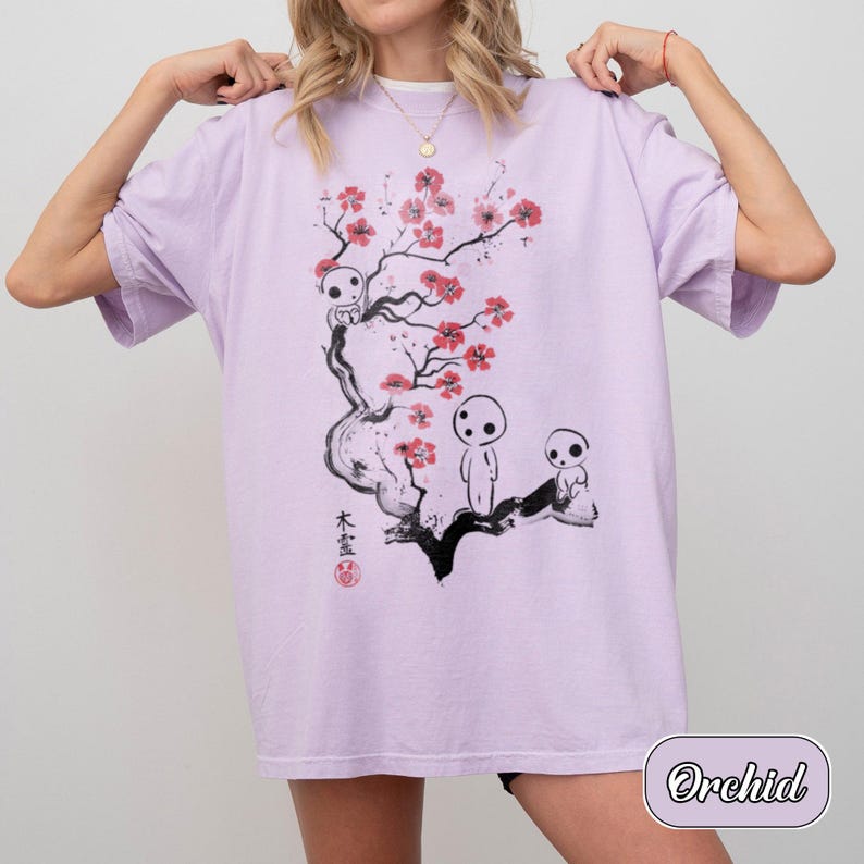 May include: A light purple t-shirt featuring a black ink-style illustration of a tree with pink blossoms and three small white forest spirits. The design includes black Japanese kanji characters and a small red circular stamp near the base.