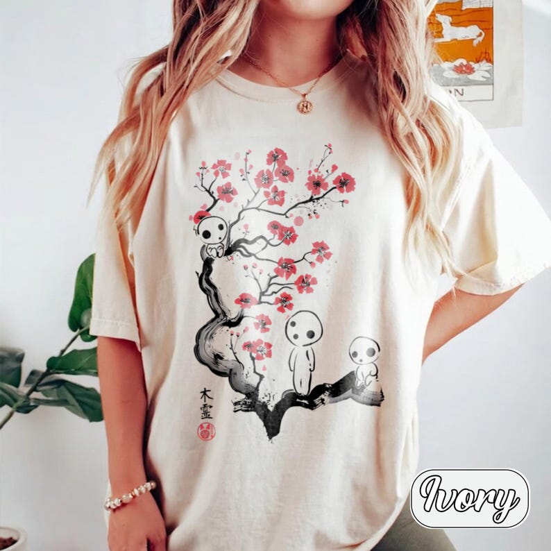May include: An ivory short-sleeve t-shirt featuring a graphic print of a dark, stylized tree with pink cherry blossoms. Three small, white, round-headed figures are positioned on the branches. Black Japanese characters are printed vertically.