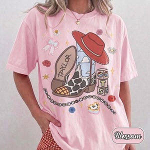Pu&ograve; includere: Una t-shirt rosa chiaro con una stampa grafica di uno stivale da cowboy con macchie di mucca e il nome TAYLOR, un cappello da cowboy rosso, blocchi con le lettere T, S e il numero 13, un fiocco, fiori e una catena di perline con testo.