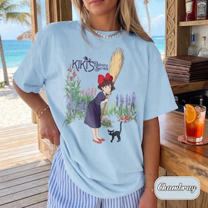 May include: A light blue t-shirt featuring a graphic print of Kiki's Delivery Service with the character Kiki holding a broomstick, a black cat, and colourful flowers. The text Kiki's Delivery Service is printed above the character illustration.