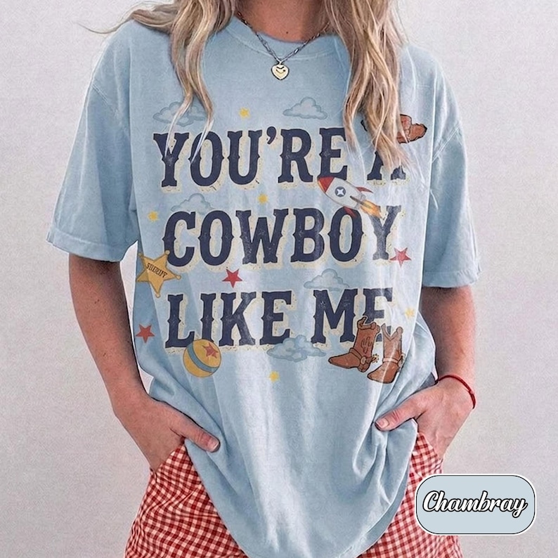 K&ouml;nnte beinhalten: Ein hellblaues Kurzarm-T-Shirt mit dem dunkelblauen Schriftzug "YOU'RE A COWBOY LIKE ME", umgeben von grafischen Illustrationen einer Rakete, eines Sheriffsterns, eines Balls, Wolken und braunen Cowboystiefeln mit kleinen Sternen.
