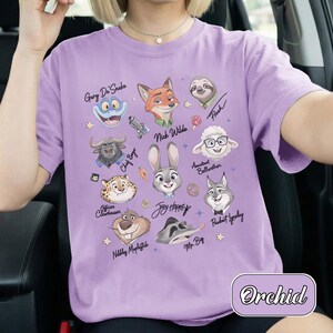 May include: A lilac t-shirt with cartoon portraits of Zootopia characters, such as Nick Wilde and Judy Hopps. Each character has their name above their portrait. The word "Orchid" is printed on the bottom right of the shirt.