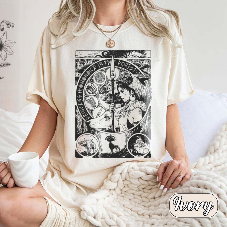 以下が含まれることがあります： アイボリーの半袖Tシャツ。槍を持つキャラクターの黒いグラフィックプリントが施され、円形のモチーフと「Princess Mononoke in the Forest」というテキストで囲まれています。デザインには様式化された森の動物や植物が含まれています。