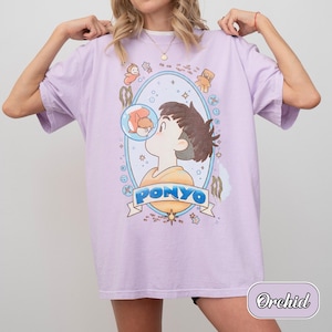 May include: An orchid-coloured t-shirt featuring a graphic print of a character and a goldfish inside a bubble. The design is framed by an oval border with small icons and the text PONYO in blue letters on a banner at the bottom of the illustration.