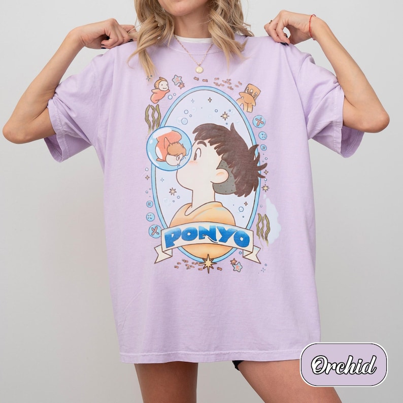 May include: An orchid-coloured t-shirt featuring a graphic print of a character and a goldfish inside a bubble. The design is framed by an oval border with small icons and the text PONYO in blue letters on a banner at the bottom of the illustration.