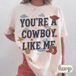 K&ouml;nnte beinhalten: Ein elfenbeinfarbenes Kurzarm-T-Shirt mit dem dunkelblauen Text "YOU'RE A COWBOY LIKE ME", umgeben von Illustrationen eines braunen Cowboyhuts, einer Rakete, eines gelben Sheriffsterns, eines bunten Balls, brauner Cowboystiefel und blauer Wolken.