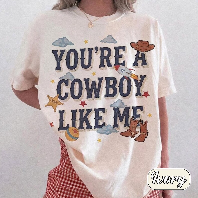 K&ouml;nnte beinhalten: Ein elfenbeinfarbenes Kurzarm-T-Shirt mit dem dunkelblauen Text "YOU'RE A COWBOY LIKE ME", umgeben von Illustrationen eines braunen Cowboyhuts, einer Rakete, eines gelben Sheriffsterns, eines bunten Balls, brauner Cowboystiefel und blauer Wolken.