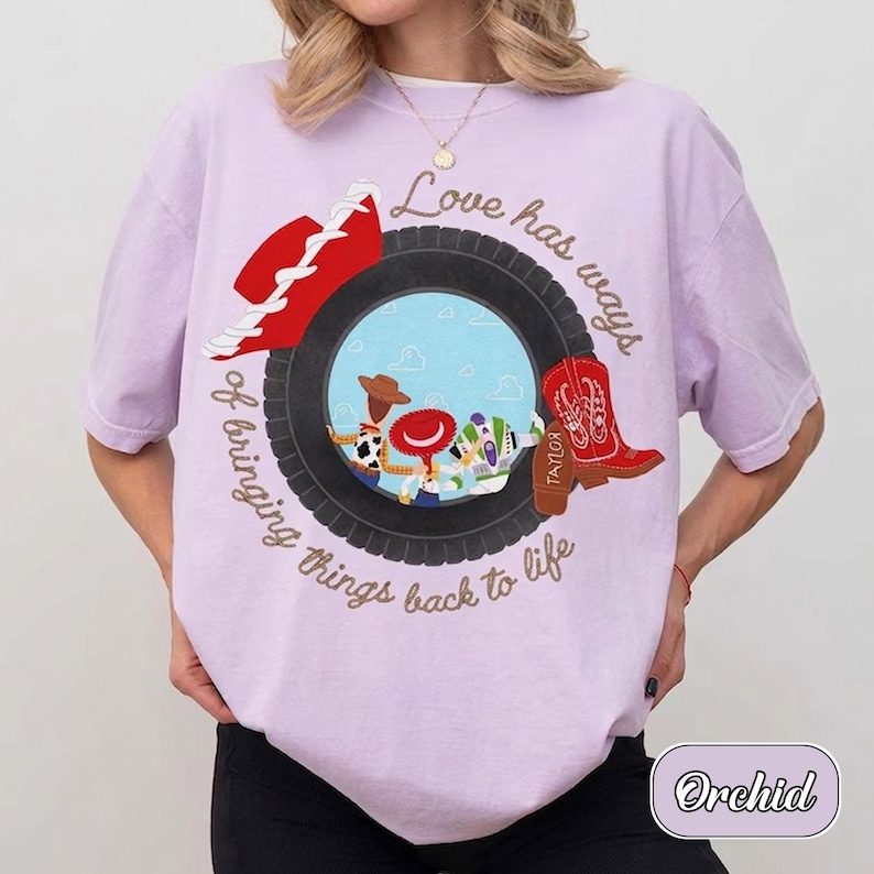 May include: A light purple t-shirt featuring a graphic of a black tire surrounding cartoon characters. The text reads Love has ways of bringing things back to life. A red cowboy hat and a red cowboy boot are positioned on the tire graphic.
