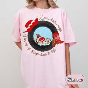 May include: A light pink t-shirt featuring a graphic of a black tire surrounding a blue sky scene with three cartoon characters. The design includes a red hat, a red cowboy boot, and the text "Love has ways of bringing things back to life."