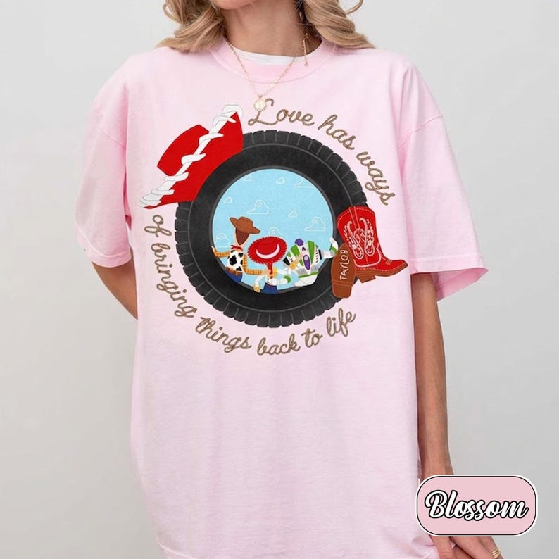 May include: A light pink t-shirt featuring a graphic of a black tire surrounding a blue sky scene with three cartoon characters. The design includes a red hat, a red cowboy boot, and the text "Love has ways of bringing things back to life."