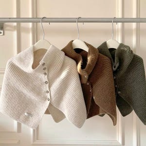 Op de afbeelding: Drie gebreide capes in crème, bruin en olijfgroen hangen aan een metalen stang. Elke cape heeft een brede kraag, een knoopsluiting en een textuur gebreid ontwerp. De capes worden gepresenteerd op witte hangers.