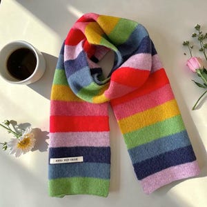 May include: A vibrant rainbow striped scarf with a label reading "ANNA WORTHIE". The scarf is made of a soft, textured material and is displayed on a white surface. A cup of coffee and flowers are also in the image.