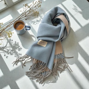 May include: A blue and beige scarf with fringed edges. The scarf is arranged in folds, with a small label attached. A cup of coffee is placed nearby on a white surface, with a sprig of dried flowers.