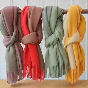 Peut inclure: Quatre écharpes colorées sont suspendues à une tringle en bois. Les écharpes sont dans des tons de mauve, de rouge, de marron, de vert sauge et de jaune. Chaque écharpe est nouée et a une bordure à franges. Le fond est un mur blanc avec des panneaux en bois verticaux.