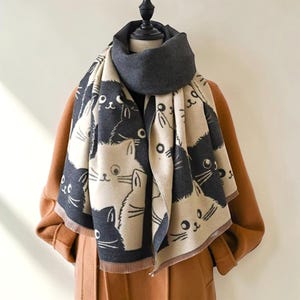 May include: A beige and charcoal cat-print scarf draped over a brown coat. The scarf features a repeating pattern of cat faces in charcoal against a beige background. A solid charcoal scarf is wrapped around the neck. The scarf is made of a soft material.