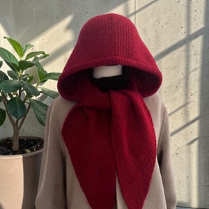 May include: A red knit hood and scarf set. The hood is a deep red color and has a wide brim. The scarf is long and drapes down the front. The set is made of a textured knit material. The background is a neutral color.
