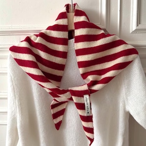 May include: A red and cream striped scarf is displayed on a white jumper. The scarf is knotted at the front. The scarf has a tag that reads "JOOM JOOM VCCM".