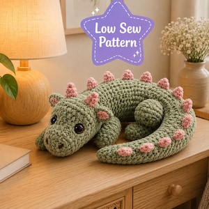 Puede incluir: Un peluche de dinosaurio tejido a ganchillo en color verde salvia con púas rosas a lo largo de la espalda y la cola. El juguete tiene ojos oscuros grandes y una superficie de hilo texturizado. Un gráfico morado muestra Low Sew Pattern.