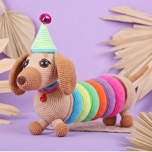May include: A crocheted dachshund toy with a tan body and long ears. The dog wears a light green and blue party hat with a pink pom-pom, a blue collar with a small red heart charm, and a colorful rainbow-striped body section in bright neon hues.