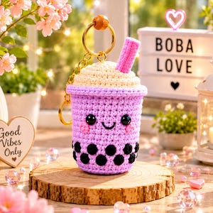 Può includere: Un portachiavi boba tea all'uncinetto nei toni del viola, crema e rosa, con un volto sorridente e perle di boba nere. Il portachiavi è su una fetta di legno, con una catena dorata e una perlina di legno. Lo sfondo include un cartello con la scritta "BOBA LOVE".
