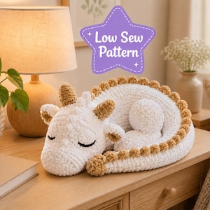 May include: A white crocheted dragon plush toy with tan horns and a tan spiked ridge along its back. The dragon is in a curled sleeping position with closed eyes. A purple star-shaped graphic above the toy contains the text Low Sew Pattern.