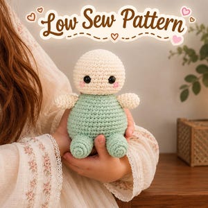 May include: A small, round, handmade crochet doll with a cream-colored head, black safety eyes, pink cheeks, and a mint green body with matching arms and legs. The text "Low Sew Pattern" is displayed in brown cursive font above the doll.