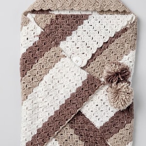 May include: A handmade crochet baby swaddle in a neutral color palette. The swaddle features a hood and a diagonal striped pattern in white, brown, and beige. It has a button closure and two decorative pom-poms. The swaddle is designed for newborns.