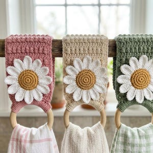 Op de afbeelding: Drie handgemaakte gehaakte keukentowelhouders in roze, beige en groen, elk versierd met een grote madelief. De madeliefjes hebben witte bloemblaadjes en een gele kern. De handdoeken hangen aan houten ringen.