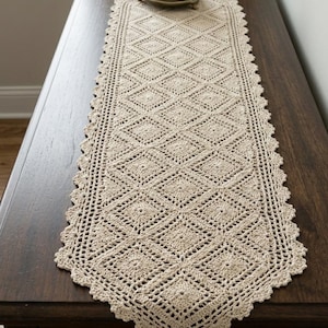 May include: A beige, handmade crochet table runner with a diamond pattern and scalloped edges. The runner is rectangular with a tassel at the end. A small potted plant sits on top of the runner.