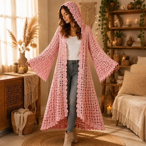 Puede incluir: Un cárdigan largo de ganchillo en color rosa claro con capucha, patrón de punto abierto, mangas acampanadas y borde festoneado. La prenda se lleva sobre una camiseta blanca lisa y vaqueros azules, llegando hasta la parte baja de la pantorrilla.