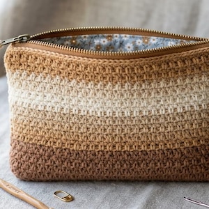 Peut inclure: Une pochette crochetée dans les tons marron, beige et crème, avec une fermeture éclair. L'intérieur est doublé d'un motif floral. Un crochet, du fil et un marqueur de point sont à côté de la pochette.