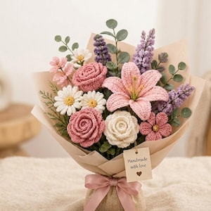 Op de afbeelding: Een gehaakt boeket met roze rozen, witte madeliefjes en een roze lelie. De bloemen zijn verpakt in bruin papier en vastgebonden met een roze lint. Een label met "Handmade with love" is bevestigd.