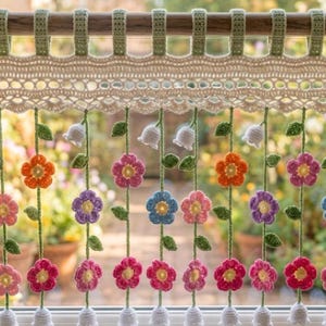 Könnte beinhalten: Ein handgefertigter Fensterbehang aus Häkelarbeit mit floralem Design. Der Behang hat eine cremefarbene Spitzenspitze mit hängenden Häkelblumen in verschiedenen Farben, darunter Rosa, Orange, Blau und Lila. Grüne Blätter und weiße glockenförmige Blumen sind ebenfalls enthalten.
