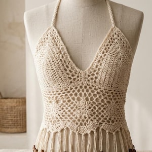 May include: Cream-colored crochet halter top with a V-neck design. The top features a detailed openwork pattern and a fringe hemline adorned with small wooden beads. The halter straps are braided. The top is displayed on a mannequin.