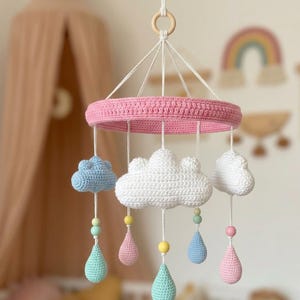 May include: A handmade baby mobile featuring a pink crocheted ring with hanging white, blue, and white crocheted clouds and teardrops. The mobile is suspended from a wooden ring and white string. Pastel colored beads accent the teardrops.