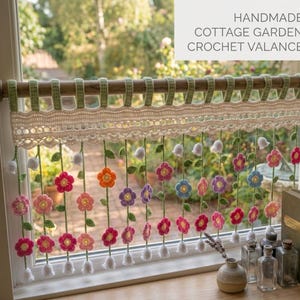 May include: A handmade crochet valance featuring a cream-colored lace header with green loops hanging from a wooden rod. Below the header, multiple green yarn stems dangle, each adorned with small, colorful crocheted flowers and white bells.