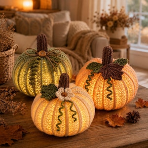May include: Three crocheted pumpkins in light green, cream, and orange, each featuring a dark brown stem. The pumpkins are illuminated from within, casting a warm glow. The green pumpkin has a small wooden button, while the cream one features a crocheted flower and green curly vines.