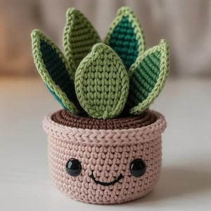 Puede incluir: Una planta suculenta de ganchillo en una maceta rosa con una cara sonriente. La planta tiene hojas verdes con detalles en verde oscuro y verde claro. La maceta es de color rosa claro con una base marrón y un borde rosa claro. Los ojos son negros y la boca es una sonrisa cosida.